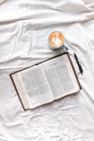 An open book displaying a page titled '2 Kings' is placed on a white fabric surface. Beside the book, there is a pen and a mug containing a latte with art on top.