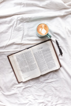 An open book displaying a page titled '2 Kings' is placed on a white fabric surface. Beside the book, there is a pen and a mug containing a latte with art on top.