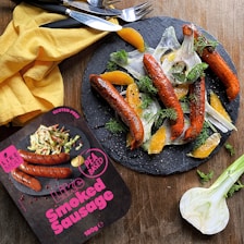 A plate with smoked sausages garnished with sliced fennel, orange segments, and fresh dill is presented on a slate serving board. Beside it, there is a yellow cloth napkin with cutlery, and a package labeled 'Like Smoked Sausage' that is pea-based and gluten-free.