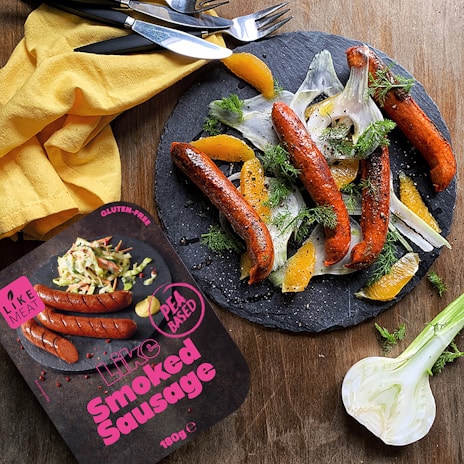 A plate with smoked sausages garnished with sliced fennel, orange segments, and fresh dill is presented on a slate serving board. Beside it, there is a yellow cloth napkin with cutlery, and a package labeled 'Like Smoked Sausage' that is pea-based and gluten-free.