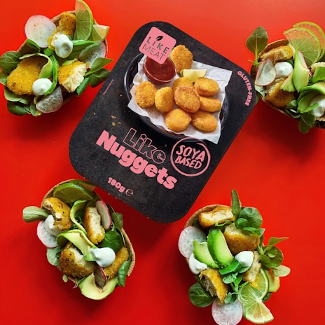 A package of plant-based nuggets is centered on a vibrant red background, surrounded by three taco-like servings filled with salad, avocado, and crispy nuggets. The package features bold text indicating the product is soya based and gluten-free.