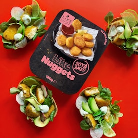 A package of plant-based nuggets is centered on a vibrant red background, surrounded by three taco-like servings filled with salad, avocado, and crispy nuggets. The package features bold text indicating the product is soya based and gluten-free.
