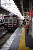 A train station platform with a maroon train arriving, passengers are waiting on the platform, some holding bags. Signs with Japanese text are prominently displayed above. The platform features a tactile yellow safety line near the edge.