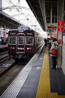A train station platform with a maroon train arriving, passengers are waiting on the platform, some holding bags. Signs with Japanese text are prominently displayed above. The platform features a tactile yellow safety line near the edge.