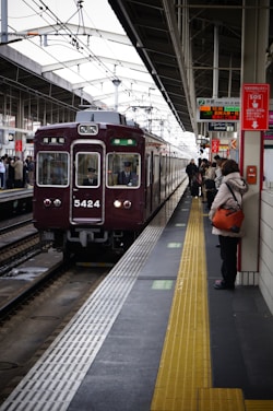 A train station platform with a maroon train arriving, passengers are waiting on the platform, some holding bags. Signs with Japanese text are prominently displayed above. The platform features a tactile yellow safety line near the edge.