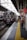 A train station platform with a maroon train arriving, passengers are waiting on the platform, some holding bags. Signs with Japanese text are prominently displayed above. The platform features a tactile yellow safety line near the edge.