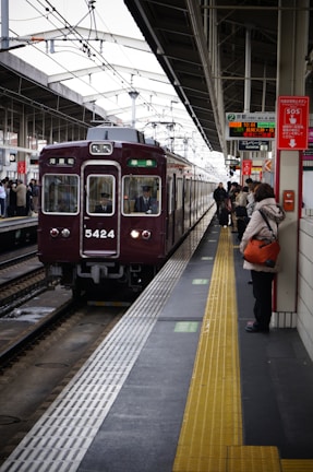 A train station platform with a maroon train arriving, passengers are waiting on the platform, some holding bags. Signs with Japanese text are prominently displayed above. The platform features a tactile yellow safety line near the edge.