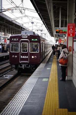 A train station platform with a maroon train arriving, passengers are waiting on the platform, some holding bags. Signs with Japanese text are prominently displayed above. The platform features a tactile yellow safety line near the edge.