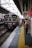 A train station platform with a maroon train arriving, passengers are waiting on the platform, some holding bags. Signs with Japanese text are prominently displayed above. The platform features a tactile yellow safety line near the edge.