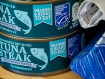 Two cans of responsibly sourced tuna steak in spring water, labeled with certifications for being drained and ready to eat and certified sustainable seafood. The cans are by Sainsbury's and feature an illustrated fish logo. The second can is partially covered by what appears to be a rolled-up piece of paper. There is a blue-striped object to the right side.