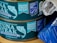 Two cans of responsibly sourced tuna steak in spring water, labeled with certifications for being drained and ready to eat and certified sustainable seafood. The cans are by Sainsbury's and feature an illustrated fish logo. The second can is partially covered by what appears to be a rolled-up piece of paper. There is a blue-striped object to the right side.