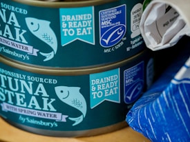 Two cans of responsibly sourced tuna steak in spring water, labeled with certifications for being drained and ready to eat and certified sustainable seafood. The cans are by Sainsbury's and feature an illustrated fish logo. The second can is partially covered by what appears to be a rolled-up piece of paper. There is a blue-striped object to the right side.