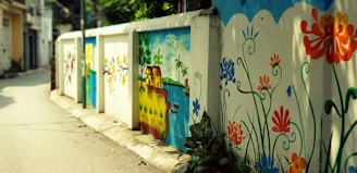 A vibrant street mural decorates a low wall with colorful floral patterns and a serene landscape scene. The mural includes orange, blue, and red flowers intertwined with green leaves, alongside an idyllic depiction of a house beside a river under a sunny sky.