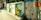 A vibrant street mural decorates a low wall with colorful floral patterns and a serene landscape scene. The mural includes orange, blue, and red flowers intertwined with green leaves, alongside an idyllic depiction of a house beside a river under a sunny sky.