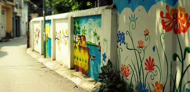 A vibrant street mural decorates a low wall with colorful floral patterns and a serene landscape scene. The mural includes orange, blue, and red flowers intertwined with green leaves, alongside an idyllic depiction of a house beside a river under a sunny sky.