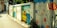 A vibrant street mural decorates a low wall with colorful floral patterns and a serene landscape scene. The mural includes orange, blue, and red flowers intertwined with green leaves, alongside an idyllic depiction of a house beside a river under a sunny sky.