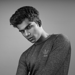 A grayscale portrait of a person with a serious expression, wearing a textured dark shirt with a logo on it. The hair is styled upward, and the head is slightly tilted. The background is plain and gray, emphasizing the subject.