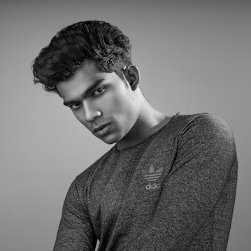 A grayscale portrait of a person with a serious expression, wearing a textured dark shirt with a logo on it. The hair is styled upward, and the head is slightly tilted. The background is plain and gray, emphasizing the subject.