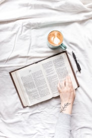 An open book is placed on a white fabric surface. A hand with a tattoo of a leaf on the wrist is gently touching the pages. Next to the book, there is a blue mug containing a cappuccino with latte art, and a black pen lies nearby.
