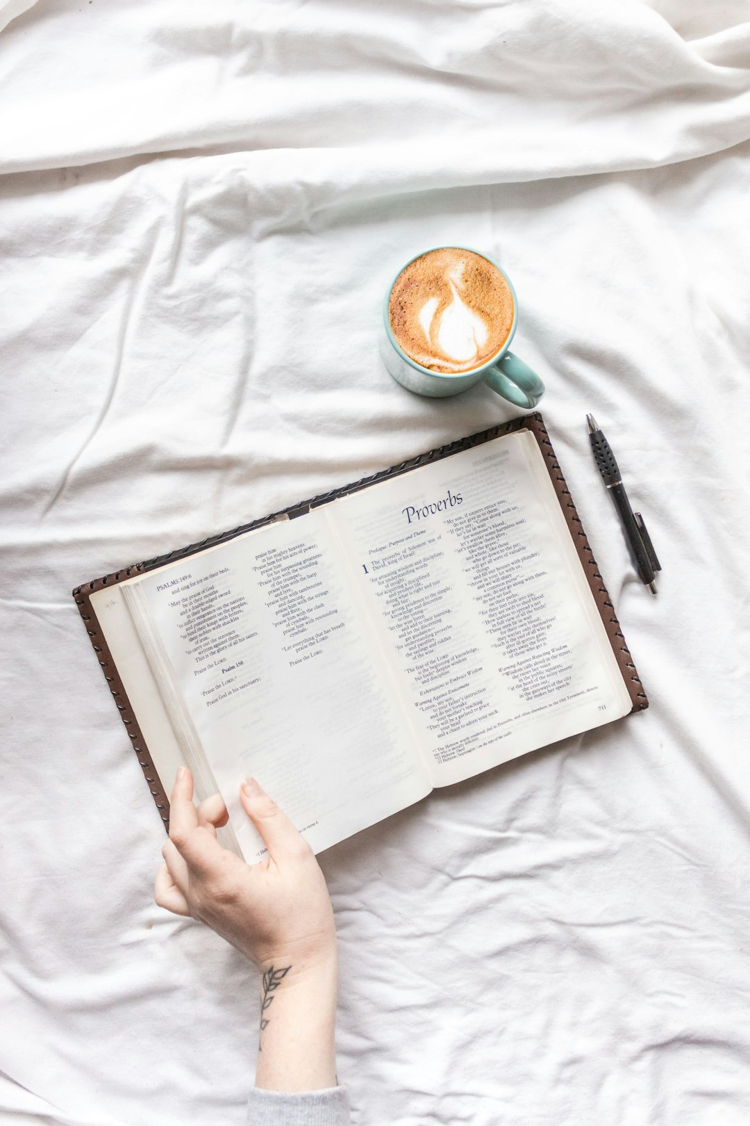 person holding white book page beside black click pen and white ceramic mug on white textile