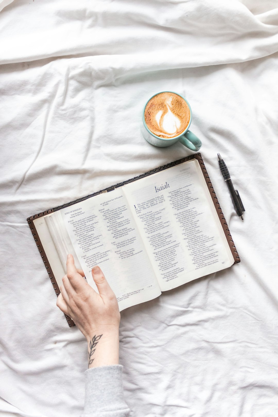 person holding white book page near black click pen and white ceramic mug on white textile