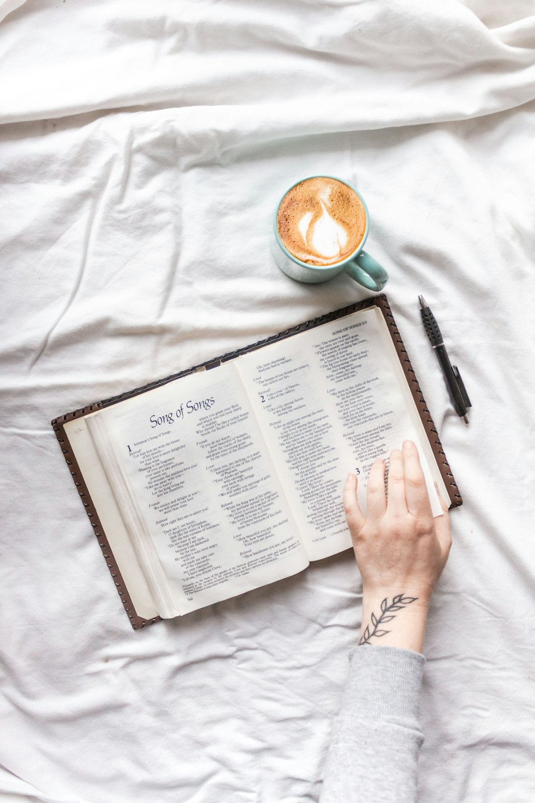 person holding white book page beside black pen and white ceramic mug on white textile