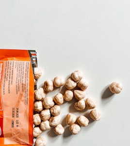 An open packet of hazelnuts spills out onto a light-colored surface. The packaging is orange with black and white text, and several whole hazelnuts are visible next to it.