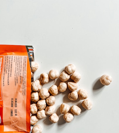 An open packet of hazelnuts spills out onto a light-colored surface. The packaging is orange with black and white text, and several whole hazelnuts are visible next to it.