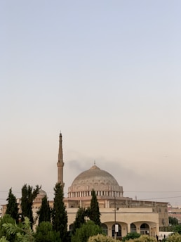 A large mosque with a prominent dome and a tall minaret stands amidst lush greenery. The architecture features classic Islamic elements with arches and detailed stonework. The sky is clear and bright, suggesting a peaceful atmosphere.