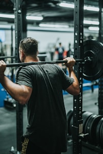 A person is performing a squat exercise with a loaded barbell on their shoulders in a gym. The setting is a well-equipped fitness facility with various weights and equipment visible in the background.