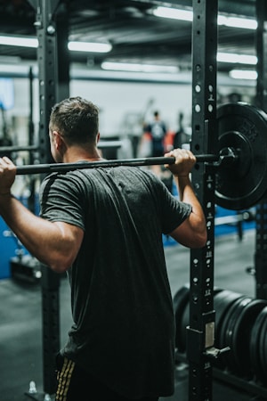 A person is performing a squat exercise with a loaded barbell on their shoulders in a gym. The setting is a well-equipped fitness facility with various weights and equipment visible in the background.