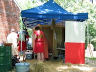 Several people dressed in historic or Roman-style costumes stand and interact under a blue canopy tent. The tent is situated outdoors with hay on the ground, and a red and white panel partially blocks the entrance. A brick building is visible on the left side, and the surrounding area is lush with green trees.