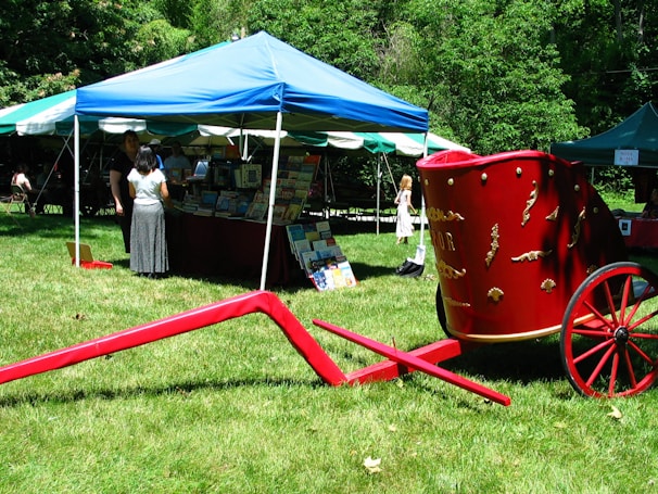 A vibrant outdoor book fair featuring a blue and white tent with book displays inside. People are browsing through the books. A striking red wagon with decorative details stands on the grass in the foreground. Trees and more tents are visible in the background.