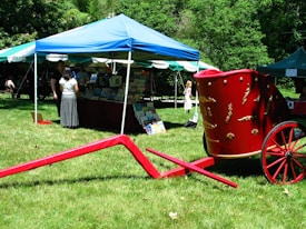 A vibrant outdoor book fair featuring a blue and white tent with book displays inside. People are browsing through the books. A striking red wagon with decorative details stands on the grass in the foreground. Trees and more tents are visible in the background.