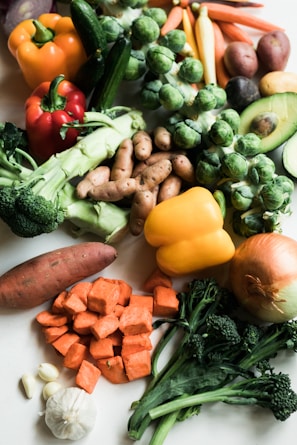 A variety of fresh vegetables are arranged on a light surface, including bell peppers, broccoli, Brussels sprouts, carrots, potatoes, an avocado, garlic, and an onion. The vegetables exhibit a range of colors and textures, creating a vibrant and healthy display.
