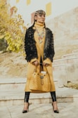 A person stands confidently on steps, wearing a long yellow turtleneck dress paired with a black fur jacket. The outfit is accessorized with a colorful beaded necklace and a large, quilted yellow handbag. The ensemble is completed with black leggings and high-heeled shoes. The background features stone steps and some autumn foliage.