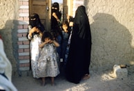 A group of women and children are gathered near a doorway in a rustic setting. The women are wearing traditional clothing with face coverings, while the children are in simple, colorful dresses. The wall to their right is made of mud bricks, and the ground is sandy. The scene is calm with natural light casting shadows.
