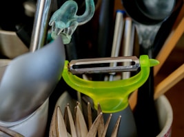 A collection of kitchen utensils is arranged in a holder. A bright green peeler is prominently positioned among other items including a spoon, a pair of tongs, and some pens.