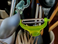 A collection of kitchen utensils is arranged in a holder. A bright green peeler is prominently positioned among other items including a spoon, a pair of tongs, and some pens.
