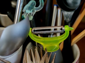 A collection of kitchen utensils is arranged in a holder. A bright green peeler is prominently positioned among other items including a spoon, a pair of tongs, and some pens.