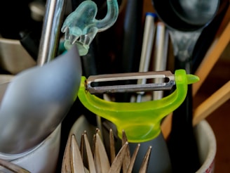 A collection of kitchen utensils is arranged in a holder. A bright green peeler is prominently positioned among other items including a spoon, a pair of tongs, and some pens.