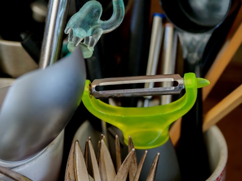 A collection of kitchen utensils is arranged in a holder. A bright green peeler is prominently positioned among other items including a spoon, a pair of tongs, and some pens.