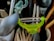 A collection of kitchen utensils is arranged in a holder. A bright green peeler is prominently positioned among other items including a spoon, a pair of tongs, and some pens.