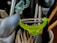 A collection of kitchen utensils is arranged in a holder. A bright green peeler is prominently positioned among other items including a spoon, a pair of tongs, and some pens.