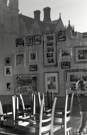 An interior scene captured in black and white, featuring an arrangement of chairs stacked upside down on tables. The walls are adorned with various framed photographs. The window reflection reveals an external building facade with chimneys and ornate architectural details.