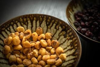 A close-up view of a bowl filled with roasted nuts, prominently displayed with a textured ceramic pattern in earthy tones, alongside another bowl containing dark dried fruits.