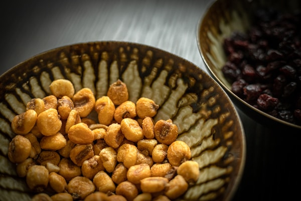 A close-up view of a bowl filled with roasted nuts, prominently displayed with a textured ceramic pattern in earthy tones, alongside another bowl containing dark dried fruits.