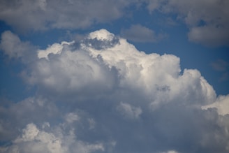 Large, fluffy clouds with varying shades of white and gray are set against a bright blue sky. The cloud formation is dense and appears to be building vertically.