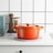 A brightly colored orange Dutch oven sits on a kitchen counter with a textured mat underneath. Nearby, there's a wooden pepper mill, a jar with walnuts, and a retro-style mint green appliance. The background features a white tiled wall with black grout.