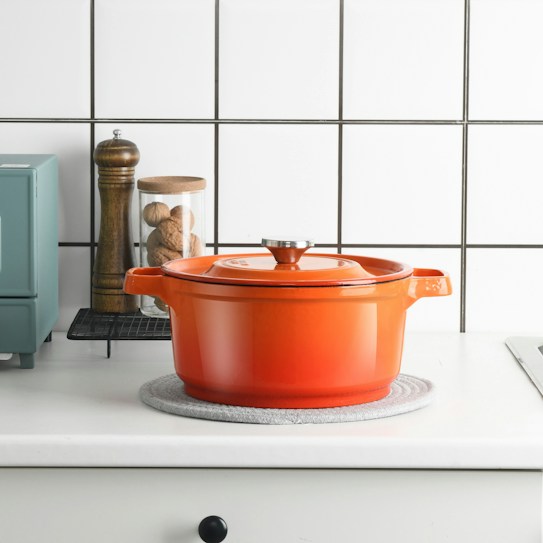 A brightly colored orange Dutch oven sits on a kitchen counter with a textured mat underneath. Nearby, there's a wooden pepper mill, a jar with walnuts, and a retro-style mint green appliance. The background features a white tiled wall with black grout.
